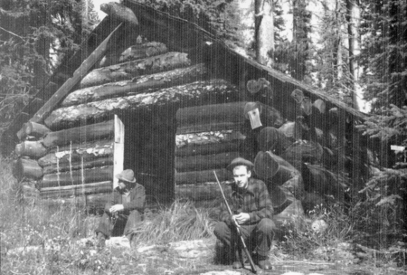 Two people sitting in front of a log cabin in the woods. One person is holding a stick, and the other person is looking to the side. The cabin is surrounded by trees and vegetation.