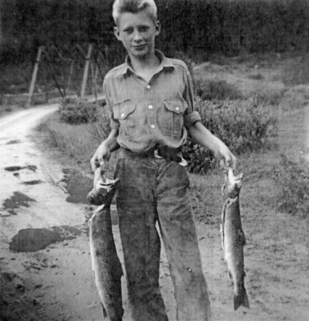 A young boy stands on a dirt path holding two fish, one in each hand. There is greenery and a bridge in the background.