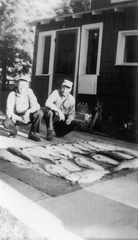 Two men are crouching on the ground next to several fish laid out in rows. They are in front of a building with a door and windows.
