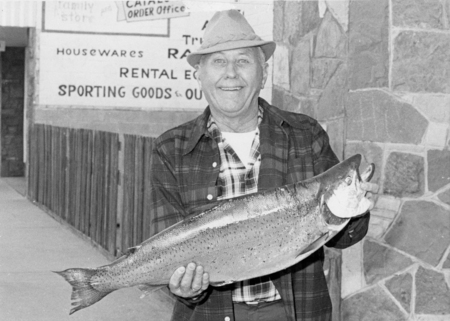 A person wearing a hat and plaid shirt is holding a large fish with a smile. They are standing in front of a stone wall and signs advertising housewares, rental equipment, and sporting goods. The visible text in the image reads: - Near the top left: "Family Store" - Near the top center: "CATAL ORDER Office" - Center left: "HOUSEWARES" - Center middle: "RENTAL EQ SPORTING GOODS & OU"
