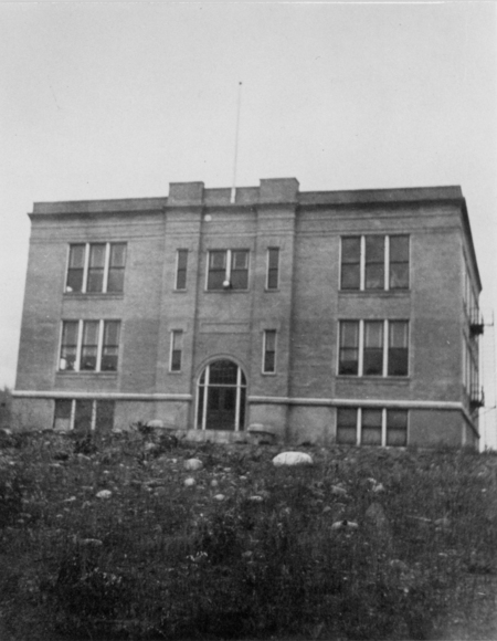 A three-story brick building with symmetrical windows and an arched entrance, situated on a grassy hill with rocks scattered in the foreground.