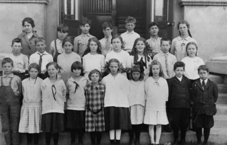 A group of children and one adult stand in front of a building. The children are arranged in rows and wear a mix of dresses, shirts, and suits, some with bows and ties. The adult is positioned at the back, slightly to the side. The setting appears to be outdoor steps leading up to a doorway.
