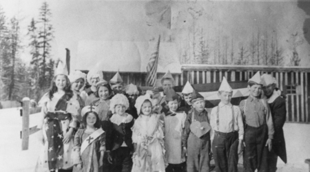 A group of children standing together outdoors, wearing various costumes and hats. Behind them are trees and a building with an American flag. Some children have capes or ribbons, and there is snow on the ground.