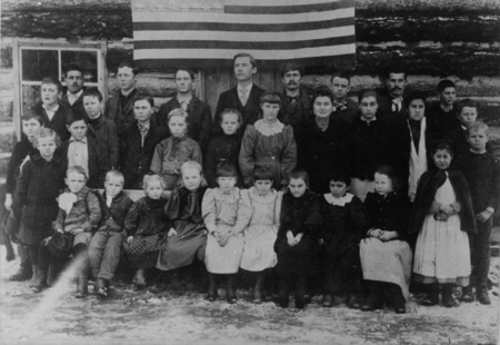 A large group of children standing in front of Priest River's first school building with an American flag draped behind them.