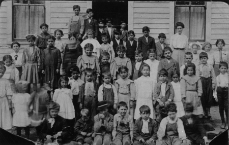 A large group of children and a woman standing in front of a wooden building. The children are arranged in several rows, with some sitting on the ground, and appear to be of varying ages. The woman stands to the side, slightly apart from the group.