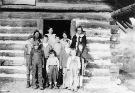 A group of children and two adults standing in front of a log cabin. The children are dressed in various attire, including overalls and hats, while the adults stand behind them at the entrance.