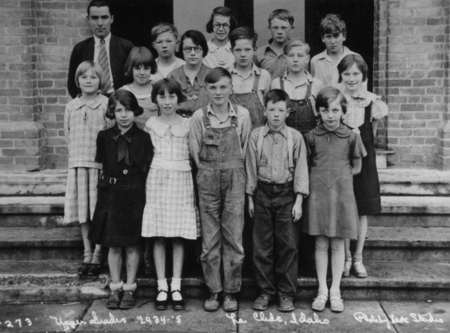 A group of children and an adult stand on the steps in front of a brick building. The children are dressed in various outfits, including dresses and overalls. The adult is wearing a suit and tie.