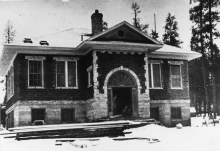A large, symmetrical brick building with a prominent arched entrance. The building has several tall windows and appears to be surrounded by snow and trees. A person or object is visible in the shadowed entranceway.