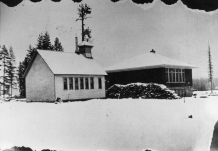 Two buildings stand in a snowy landscape. The left building has a steeple and a row of windows along its side. The right building has a flat roof and a large window in the middle. Snow covers the ground around the structures, and trees are visible in the background.