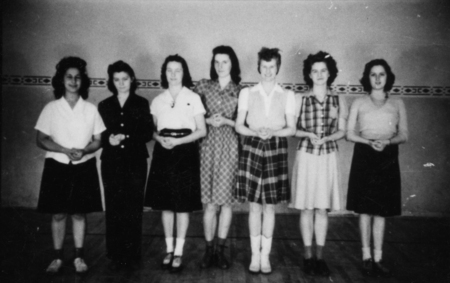 Seven women standing in a row, each with hands lightly clasped in front of them. They are wearing a variety of dresses and skirts, with different patterned and plain tops. The background has a decorative trim along the wall.