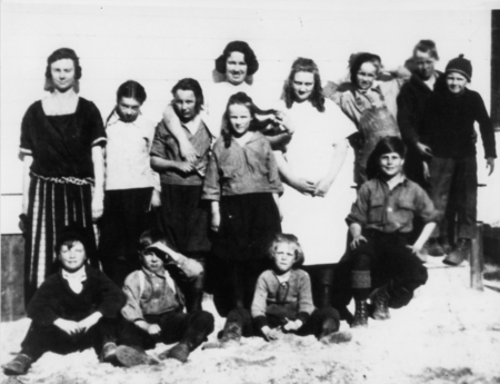 A group of twelve children and one adult standing outside in front of a building. Some children are seated on the ground, while others stand closely together. Everyone is dressed in simple clothing, and they appear to be posing for the picture.