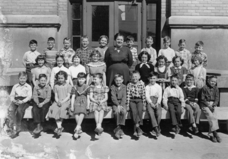 A group of young children are seated and standing in rows, with a woman standing among them, in front of a building entrance. The children are wearing a mix of dresses, shirts, and pants. The woman is wearing a dress and standing in the middle of the group.