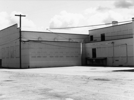 A large, flat-roofed building with few windows and a slightly weathered exterior. Power lines run across the scene, and a utility pole stands near the building. The foreground is an empty lot with a dirt surface.