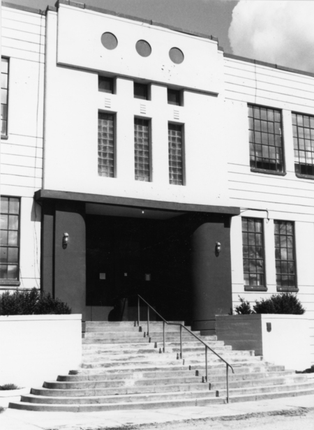 A building entrance with a series of steps leading up to double doors. The facade features three circular windows above three vertical rectangular windows. There are railings on the steps and bushes on either side of the entrance.