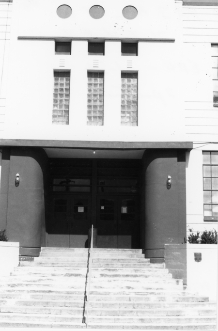 The image shows the entrance of a building with wide steps leading up to a set of double doors. Above the doors are glass block windows, and there are circular windows near the top of the building. Two sconces are mounted on the walls beside the entrance.