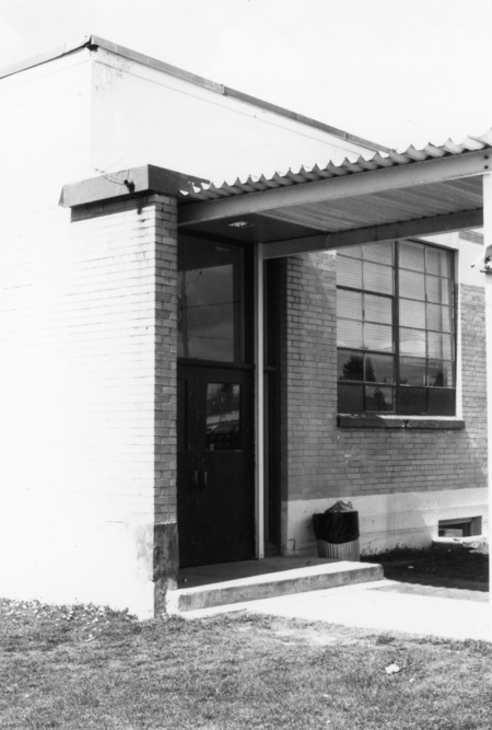 A building entrance with a covered walkway, featuring a door and a large window. A trash can is placed near the entrance on a concrete step. The building has a brick exterior and is surrounded by grass.