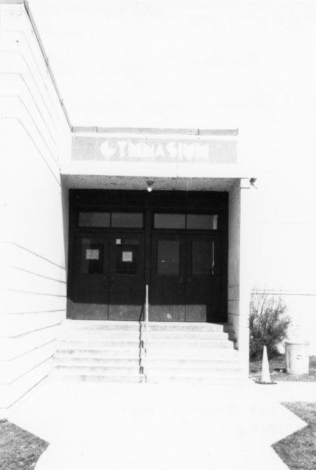 The image shows the entrance of a building labeled as a gymnasium. There are double doors with steps leading up to them. A railing runs up the center of the steps. A caution cone and a trash can are visible on one side near some bushes.