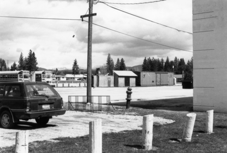 A parking lot with a car, several school buses, and some small buildings in the background. There is a utility pole and a fire hydrant nearby, with trees and mountains in the distance. The text "SCHOOL BUS" is visible near the top of two buses in the background.