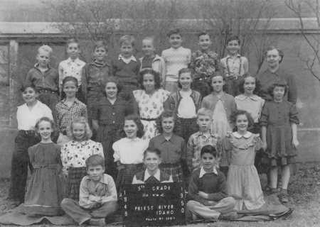 A group of children and one adult are arranged in four rows. The front row has four children sitting with a sign that reads "5th Grade 1949-1950 Priest River Idaho." Behind them stand two rows of children, with an adult on the far right side. The children and adult are dressed in various outfits, and several trees are visible in the background. Text reads: "5th GRADE 82-62 1 9 4 9 1 9 5 0 PRIEST RIVER IDAHO Photo by LEO'S".