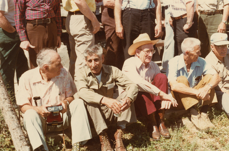 A group of seated men outdoors, with others standing behind them. One man holds a camera, another wears a cowboy hat, and some are dressed in casual shirts and slacks. The setting features grass and a wooden log.