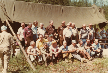 A group of men gathered outdoors, some standing and others sitting on the ground in front of a makeshift tent structure. A man is walking away from the group holding a camera in his hand. Trees are visible in the background.