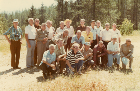 A group of men is gathered outdoors on a dirt clearing with trees in the background. Some are standing, while others are crouching in front. They wear casual clothing, and a few have name tags. One man is holding a camera.