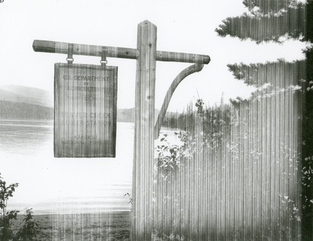 A wooden sign hanging from a wooden post near a body of water with trees and shrubs nearby. In the background, there are mountains and a forest.