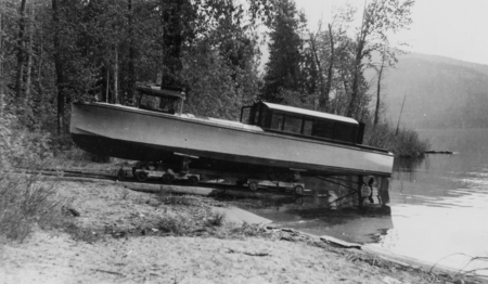 A boat is positioned on a wheeled platform near the shoreline of a lake. Trees and hills are visible in the background.
