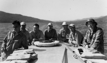 Seven men are sitting and standing on a boat, with life vests and a life preserver on the deck. They are wearing jackets and hats. The background shows water and distant hills.