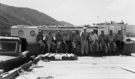 A group of men sitting on the edge of a large boat, with mountainous terrain in the background. They are dressed in casual work attire and appear to be engaging in conversation or resting. Some hold items in their hands, and equipment is visible on the deck around them.