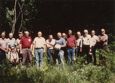 A group of thirteen men standing in a grassy area surrounded by trees. Some are wearing plaid shirts and jeans, while one man holds a hat. They are all facing the camera.