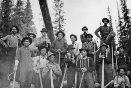 A group of men dressed in work clothes and hats standing in a forested area, holding tools such as shovels and pickaxes. They look toward the viewer with trees visible in the background.