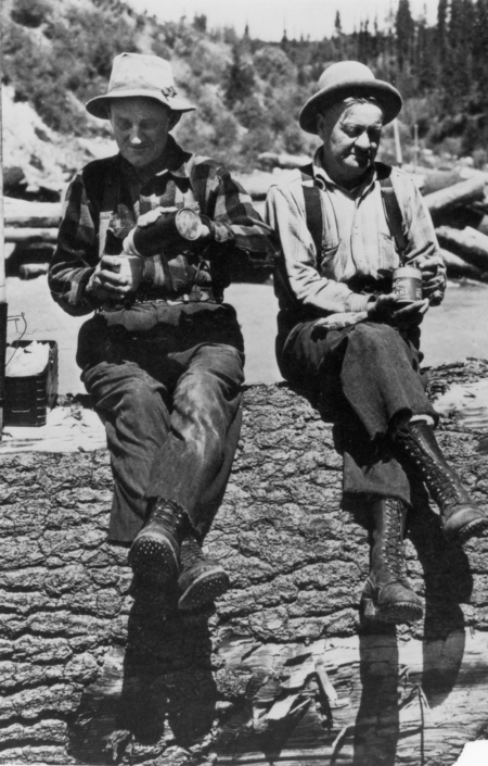 Two people sit side by side on a large log. They are both wearing hats and boots. The person on the left is pouring a drink from a thermos into a cup, while the person on the right holds a can. The background shows a natural setting with trees and rocks.