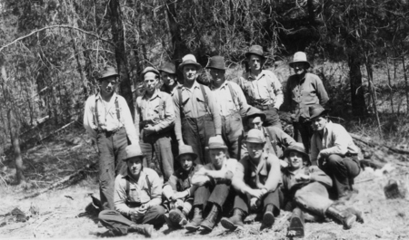 A group of men in work clothing and hats gathered together in a forested area, with some standing and others sitting or crouching on the ground. The background consists of trees and natural vegetation.