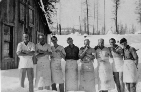 A group of eight men wearing aprons standing together outside a wooden building, with a snowy landscape and tall trees in the background.