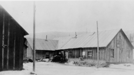 A group of wooden buildings with sloped roofs and small windows. There are a few utility poles in the foreground, and the ground is covered in snow.