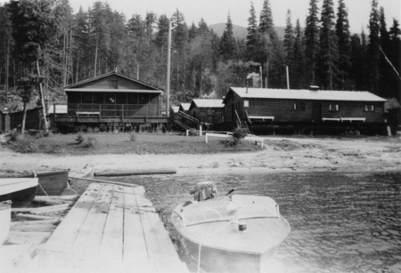 Two wooden buildings stand near a forested area with tall trees. In the foreground, a dock extends into the water, where a small boat is moored. The scene is calm with a natural, rustic atmosphere.