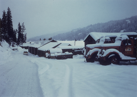 Snow-covered landscape with a road and buildings. Several trucks are parked, also covered in snow. Tall evergreen trees line the hillside on the left, and snow-capped hills are visible in the background.