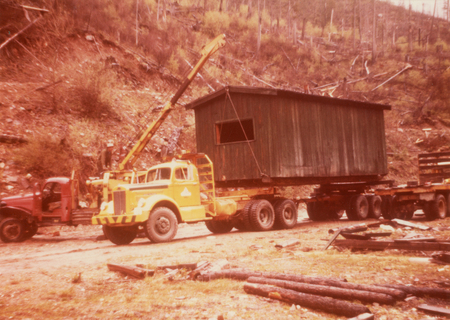 A large truck with a crane is lifting a small building onto a flatbed trailer. A person stands near the crane, and another vehicle is nearby. The area is outdoors, surrounded by rough terrain and scattered logs.
