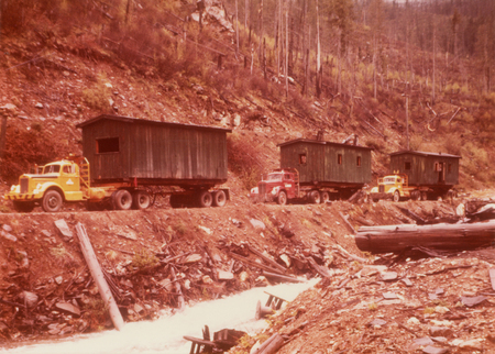 Three trucks are parked in a line on a dirt path. Each truck is carrying a large, dark green cabin-like structure on its trailer. The area around them is a rocky, wooded hillside with sparse trees and debris scattered along a nearby creek.