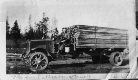 A man sitting in the driver's seat of a vintage truck loaded with lumber. The truck is parked on a dirt road with trees in the background.