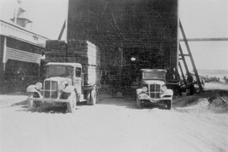 Two vintage trucks are parked in front of a large warehouse. One truck on the left is loaded with stacks of wooden crates. There are additional vehicles and a structure in the background.