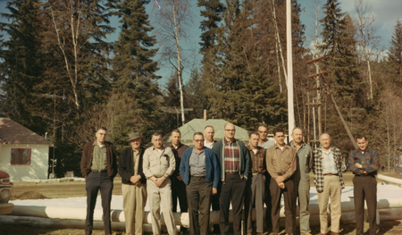 A group of twelve men standing together outdoors in front of a forest. Some are wearing jackets and hats, and a house with a green roof is visible in the background.