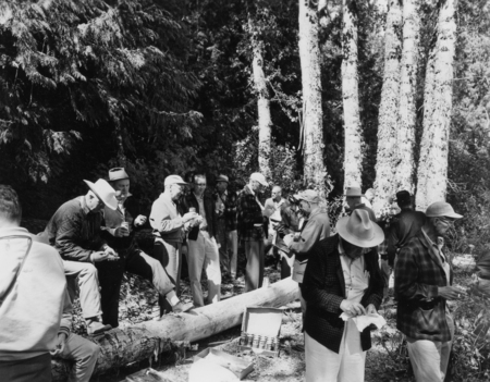 A group of people gathered in a forested area, eating and drinking a half rack of Olympia beer. They are standing and sitting on fallen logs, surrounded by tall trees and shrubs.