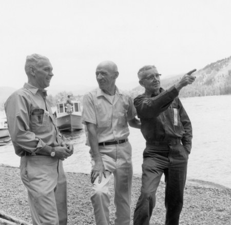 Three men standing together on a pebbled shore by the water. One man is pointing into the distance. There are boats and trees in the background.