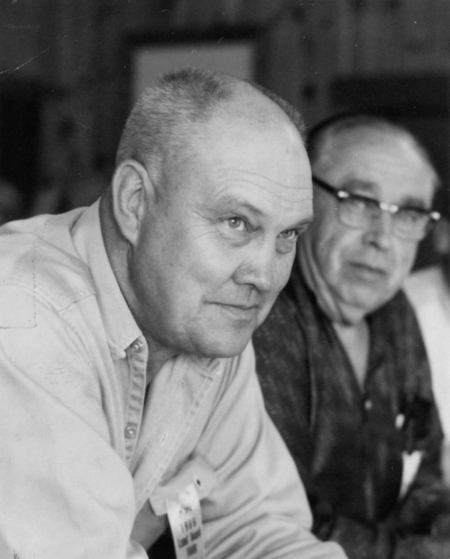 Two men sitting side by side, one in focus and the other slightly blurred. The man in focus has short hair and is wearing a button-up shirt with a name tag. The other man is wearing glasses and a patterned shirt. One wears a pendant which reads: "PLTPA (Priest Lake Timber Protection Association), 1966 Land Board Tour".