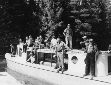 A group of men is gathered on a boat in a forested area. Some are sitting on the roof, while others are standing on the deck. They are dressed in casual outdoor clothing, and the surrounding area is filled with tall trees.