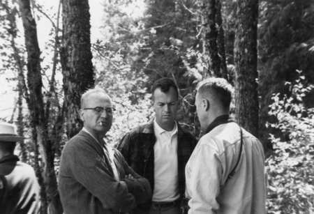 Three men stand in a wooded area. The man on the left has glasses and a cigarette in his mouth. The man in the center is looking down with a serious expression. The man on the right has his back partially to the camera. Trees and foliage surround them.