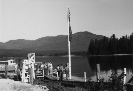 Several people stand on a dock beside a lake. A boat is moored nearby. Two flags are raised on a flagpole. In the background, there are trees and a range of hills or mountains.