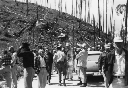A group of people, mostly men, gathered on a dirt road near a hillside with sparse, burned trees. Some wear hats and are engaged in conversation next to a car.